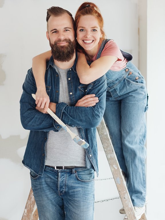 Lächelndes, liebevolles, glückliches Paar, das Renovierungsarbeiten an seiner Hoffnung durchführt und in enger Umarmung mit der jungen Frau posiert, die einen Pinsel auf einer Leiter hält.